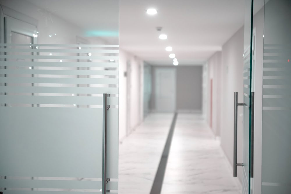 A bright, modern hallway with glass doors featuring frosted stripes and sleek metal handles, designed by a renowned commercial glass company in Union County, NJ. The corridor is lined with white walls and a marble floor, illuminated by overhead lights.