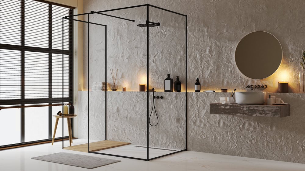 A modern bathroom with a glass shower enclosure crafted by a renowned glass contractor in Union County, NJ. Warm lighting highlights a stone-textured wall with shelves holding toiletries. A round mirror and a small wooden stool add to the minimalist decor.