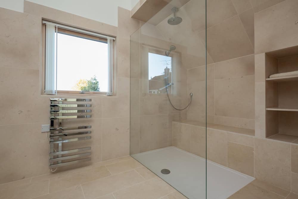 Modern bathroom with beige stone tiles, a walk-in shower installed by a premier glass contractor in Union County, NJ, and built-in shelves. A window with blinds and a sleek, metal towel radiator are on the left wall. The room is well-lit with natural light.