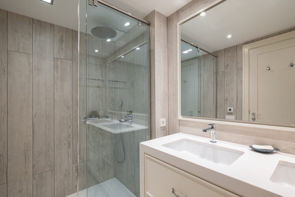 A modern bathroom featuring a glass-enclosed shower with a rain showerhead showcases the work of a renowned glass contractor in Union County, NJ. A large mirror hangs above the white double-sink vanity. Light wood paneling and recessed lighting beautifully illuminate the space.