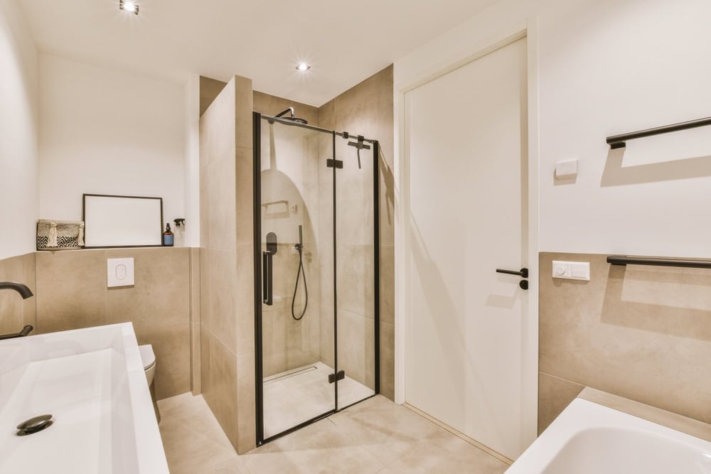 A modern bathroom designed by a renowned glass contractor in Union County, NJ, features a glass shower enclosure with a sleek black frame. Complete with a white bathtub, toilet, and sink under a mirror, the room boasts light beige tiles and a white door accented by a black handle.